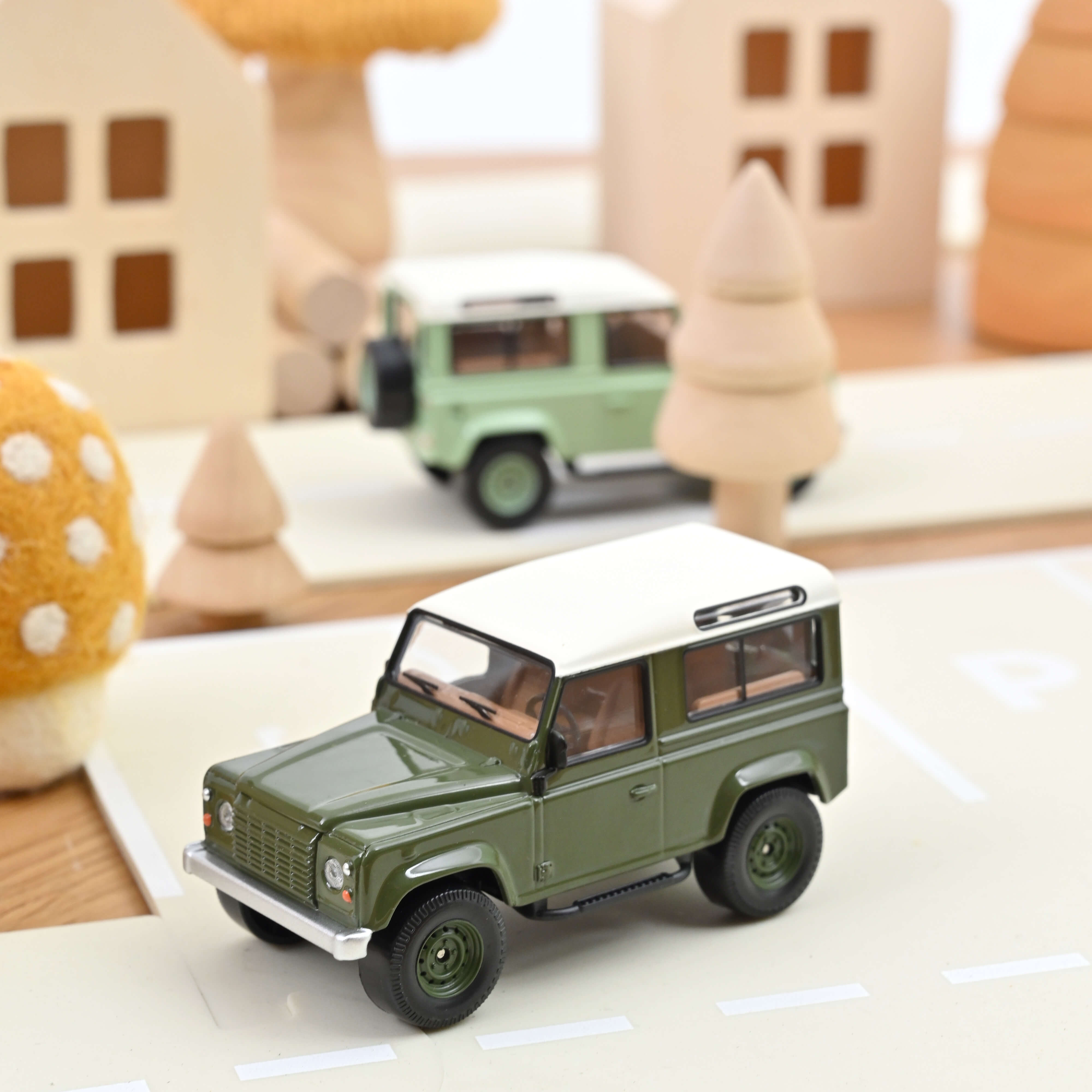 Miniature Norev Land Rover Defender 90 vert olive avec toit blanc, échelle 1/43, sur un décor de ville en bois.